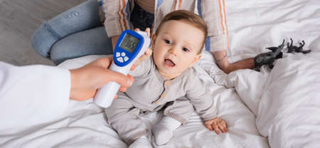 pediatrician examining infant boy with non-contact thermometer near mother, banner