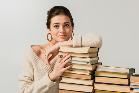 pleased young woman in earrings leaning on stack of books isolated on whiteの写真素材