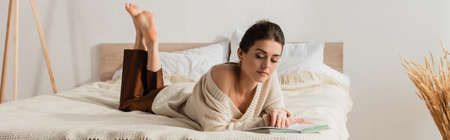 barefoot young woman reading book while resting on bed, bannerの写真素材