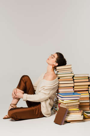 full length of young woman in glasses and sweater leaning on stack of books on whiteの写真素材