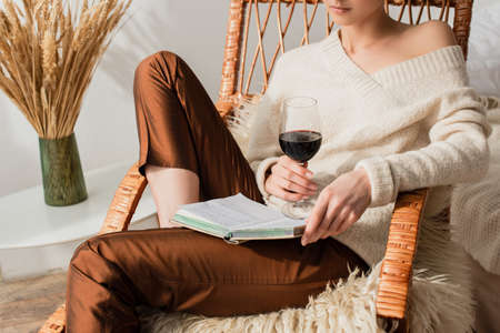 cropped view of young woman sitting in rocking chair with book and glass of wineの写真素材