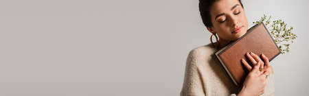 young woman with closed eyes holding book with wildflowers isolated on gray, bannerの写真素材