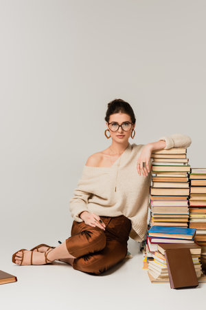 full length of young woman in glasses and sweater leaning on pile of books on whiteの写真素材