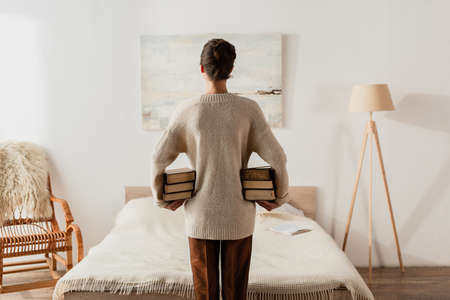 back view of woman holding stack of books in bedroomの写真素材
