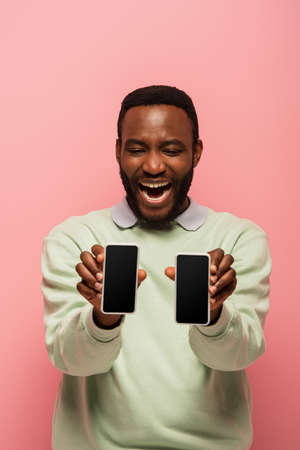 Positive african american man holding smartphones on blurred foreground isolated on pink