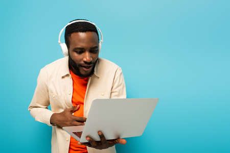 amazed african american man in headphones looking at laptop isolated on blueの写真素材