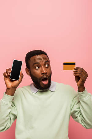 Shocked african american man looking at credit card and holding smartphone isolated on pinkの写真素材