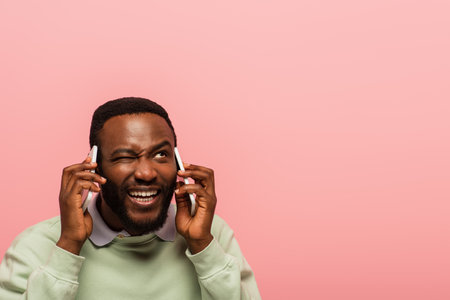 African american man winking while talking on smartphones isolated on pinkの写真素材