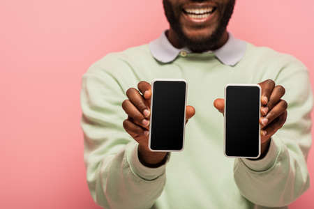 Cropped view of smartphones with blank screen in hands of smiling african american man isolated on pinkの写真素材