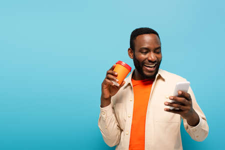 cheerful african american man with coffee to go using cellphone isolated on blue