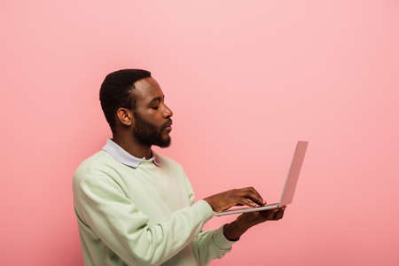 concentrated african american man typing on laptop isolated on pink