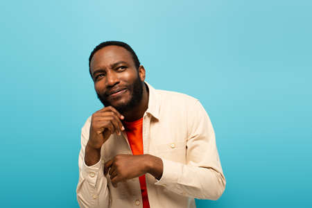 tricky african american man holding hand near face while looking away isolated on blueの写真素材