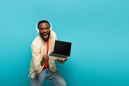 excited african american man in headphones holding laptop and showing yeah gesture on blue...