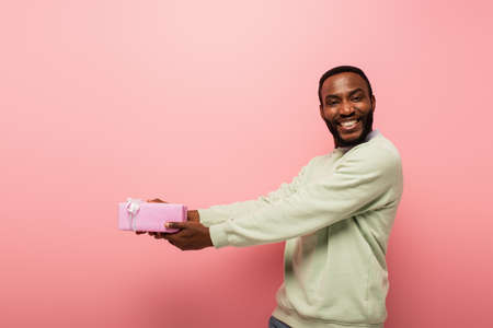 cheerful african american man holding gift in outstretched hands on pink background