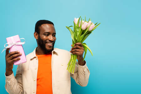 joyful african american man holding gift box and tulips isolated on blueの写真素材