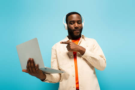 thoughtful african american man in headphones pointing at laptop on blue backgroundの写真素材
