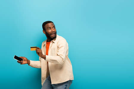 scared african american man with mobile phone and credit card looking away on blueの写真素材