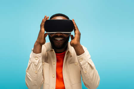 cheerful african american man adjusting vr headset while gaming isolated on blueの写真素材