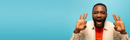 thrilled african american man showing okay signs isolated on blue, bannerの写真素材