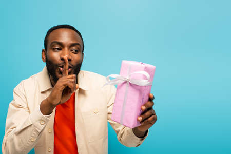 young african american man showing secret sign while holding gift box isolated on pinkの写真素材