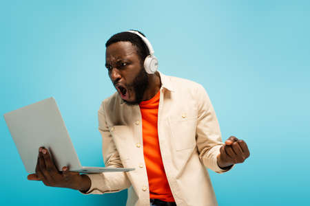 angry african american man in headphones showing clenched fist while looking at laptop on blueの写真素材