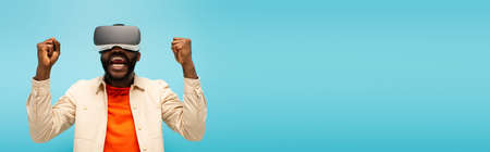 excited african american man showing win gesture while gaming in vr headset isolated on blue, banner