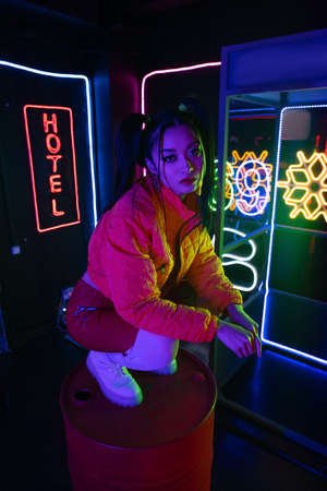 stylish young asian woman sitting near neon sign with hotel lettering outsideの写真素材