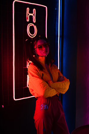 young asian woman in sunglasses standing with crossed arms near red neon hotel signの写真素材