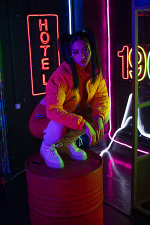 brunette asian woman sitting near neon sign with hotel lettering outsideの写真素材
