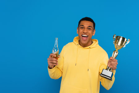 smiling african american man holding trophy cup and beer bottle in hands isolated on blue