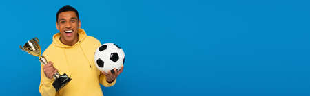 positive african american man holding football and cup in hands isolated on blue, bannerの写真素材