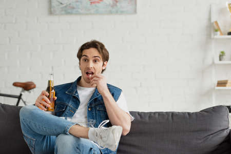 young amazed man sitting with beer and eating popcorn on couch at home