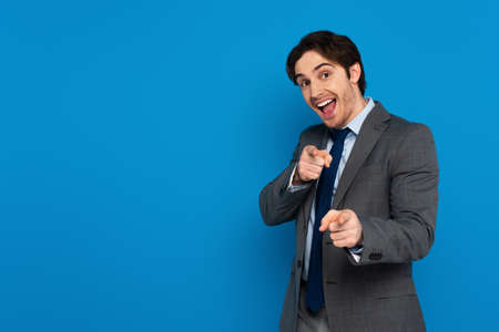 Joyful young man in elegant suit pointing with fingers at camera with smile isolated on blueの写真素材