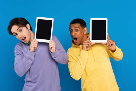 interracial friends with joyful faces holding digital devices near faces with blank screen isolated...