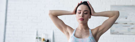 brunette woman in sports bra touching head while standing at home, bannerの写真素材