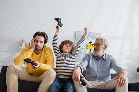 KYIV, UKRAINE - APRIL 12, 2021: Low angle view of excited kid with joystick near sad parents on couchの写真素材