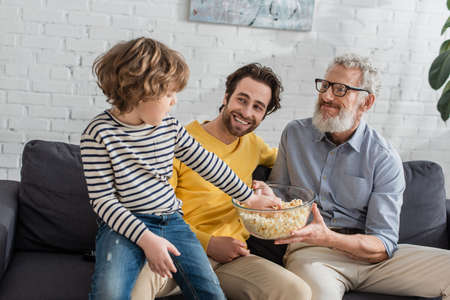 Kid taking popcorn near smiling father and grandfather at homeの写真素材