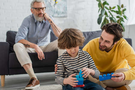 Smiling man holding building block near son and blurred father at home