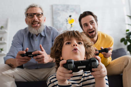 KYIV, UKRAINE - APRIL 12, 2021: Low angle view of excited child playing video game near father and granddad on blurred backgroundの写真素材