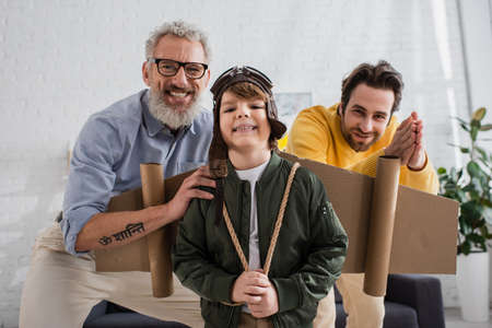Smiling kid in aviator costume looking at camera near grandfather and father