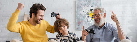 KYIV, UKRAINE - APRIL 12, 2021: Excited parents with joysticks sitting near sad kid, bannerの写真素材