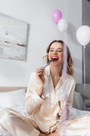 Positive woman with glass of champagne eating raspberry near balloons in bedroomの写真素材