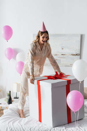 Excited woman in party cap looking at huge gift box near balloons in bedroom