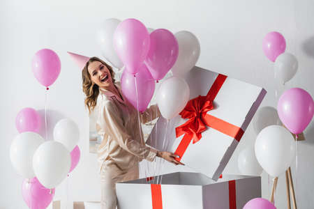 Positive woman standing near big gift box with balloons during birthday celebration at homeの写真素材