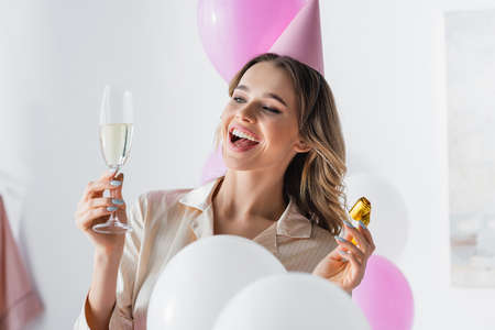 Smiling woman in party cap holding horn and champagne near balloons at homeの写真素材