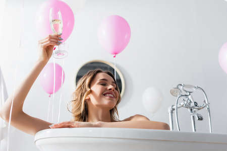Low angle view of smiling woman with glass of champagne sitting in bathtub near festive balloonsの写真素材