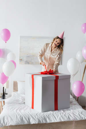 Cheerful woman in party cap holding bow of huge present near balloons in bedroomの写真素材