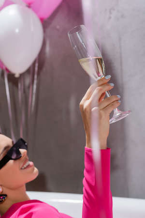Glass of champagne in hand of smiling woman in sunglasses in bathtub near balloons on blurred backgroundの写真素材