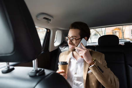 happy man in suit and glasses holding paper cup and talking smartphone in car