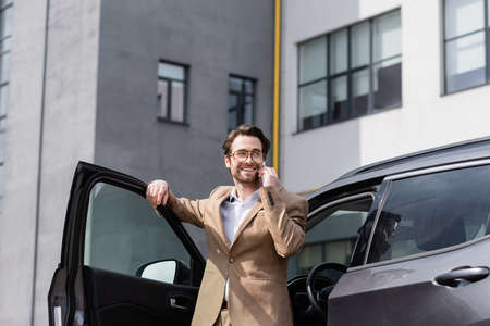 cheerful man in suit and glasses talking on smartphone and standing near car with opened doorの写真素材